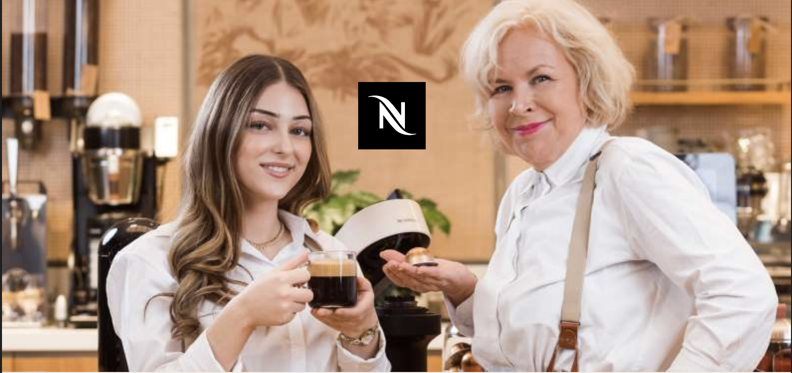 Jüngere Frau hält eine Tasse Kaffee während eine ältere blonde Frau eine Nespresso Kapsel zeigt im Hintergrund ist eine Kaffeemaschine und das Nespresso Logo.