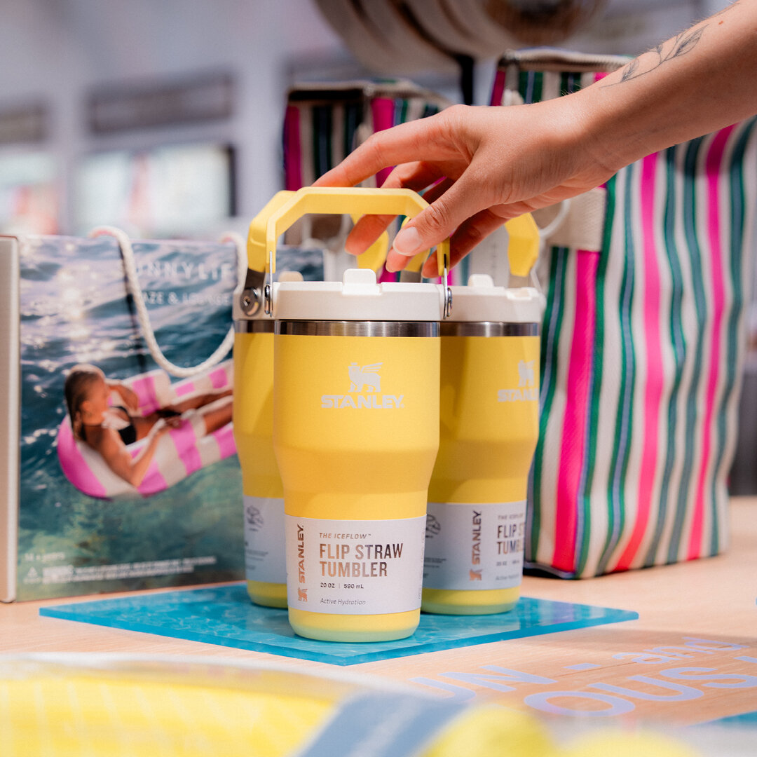 Hand greift nach einem gelben Stanley IceFlow Flip Straw Tumbler mit Tragegriff, im Hintergrund bunte Sommer-Taschen und Strandzubehör.