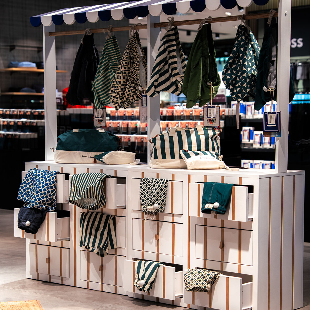 Weißer Verkaufsstand im Maritim-Look mit blau weißer Markise aufgehängten Badehosen Taschen und offenen Schubladen voller Swimwear.