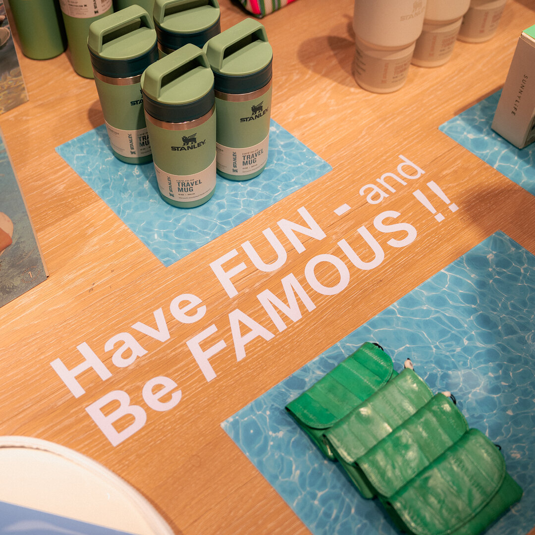 Grüne Stanley Travel Mug Isolierbecher und grüne Etuis auf Holztisch präsentiert mit weißem Schriftzug Have FUN and Be FAMOUS und blauem Wasser-Untergrund.