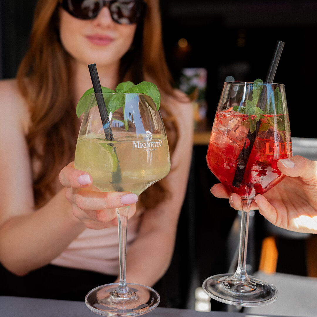 Zwei Hände stoßen mit Mionetto Prosecco und rotem Lillet Spritz an serviert im Weinglas mit Eiswürfeln Minze und Strohhalmen.