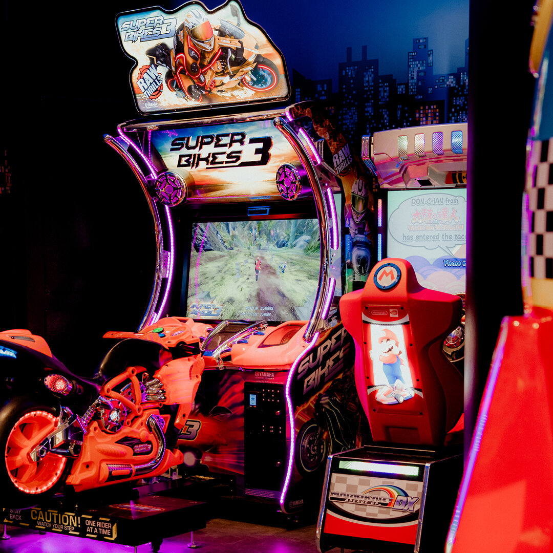 Beleuchteter Super Bikes 3 Arcade Rennspiel Automat mit orangefarbenem Motorrad zum Aufsitzen neben einem Mario Kart GP DX Automaten.