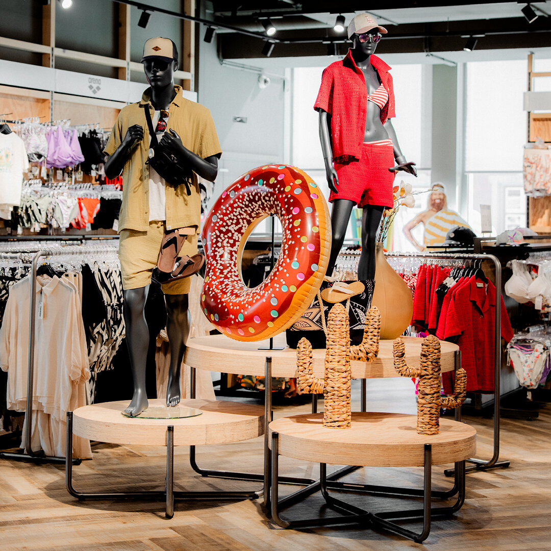 Schaufensterpuppen in gelbem und rotem Sommeroutfit auf Podesten dekoriert mit einem großen Donut Schwimmring und Flechtkakteen.