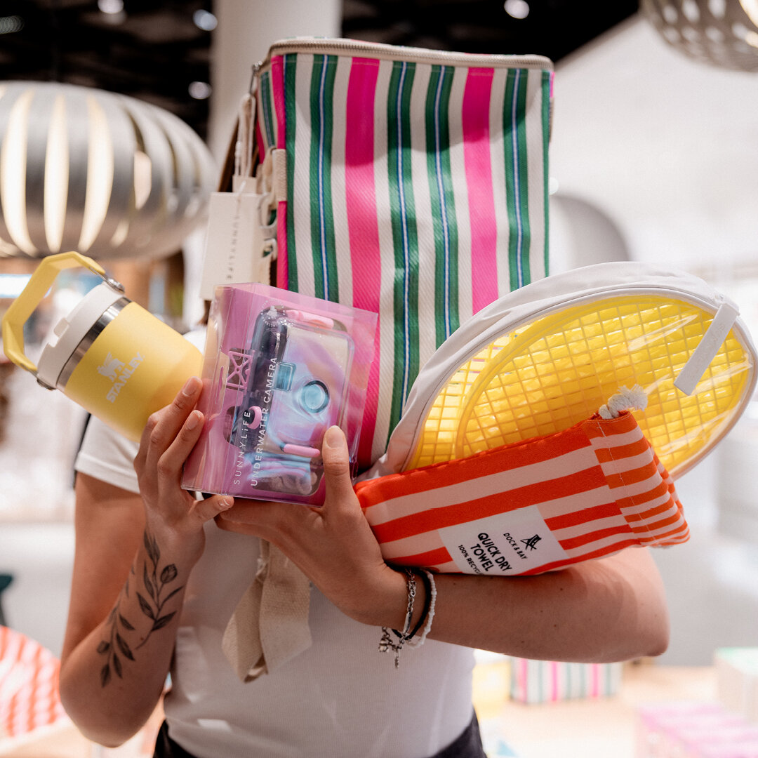 Person hält ein buntes Set mit Sommerzubehör wie gestreifte Kühltasche gelben Stanley Becher rosa Sunnylife Unterwasserkamera und Dock & Bay Strandtuch im Arm.