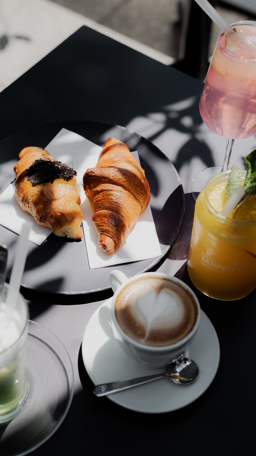 Tasse Cappuccino mit Schaumherz zwei Croissants auf einem Teller ein gelber Orangensaft und ein rosa Drink mit Eiswürfeln auf einem dunklen Café-Tisch.