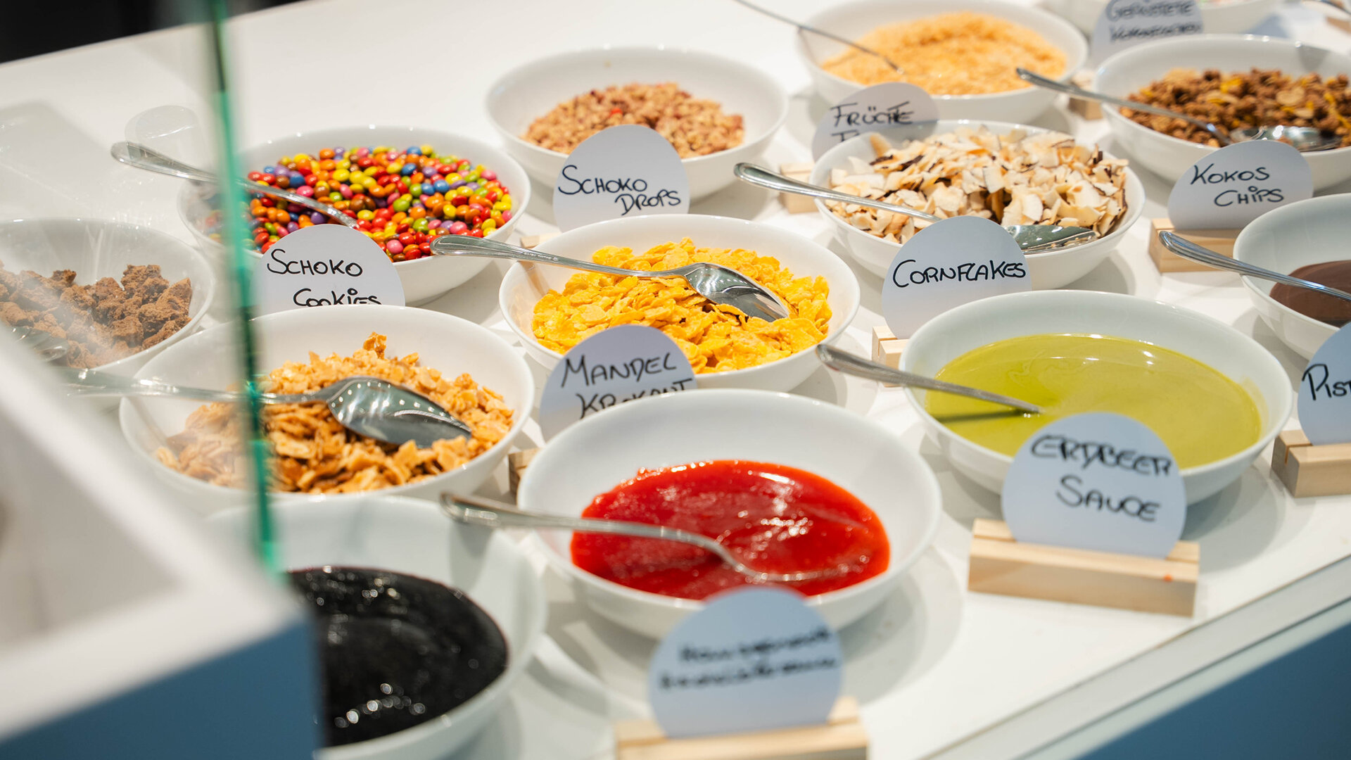 Topping-Buffet mit Schalen voller Erdbeersauce Pistaziencreme Cornflakes Schokodrops bunten Linsen und Krokant beschriftet mit kleinen Schildern.
