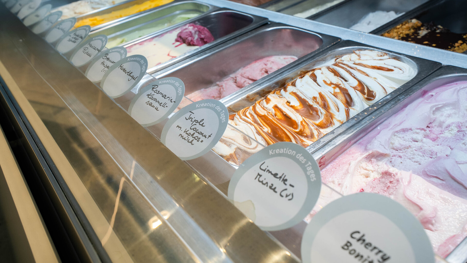 Blick in eine Eistheke mit Edelstahlbehältern voller Eissorten wie Limette-Minze und Rosmarin-Karamellsauce beschriftet mit Kreation des Tages Schildern.