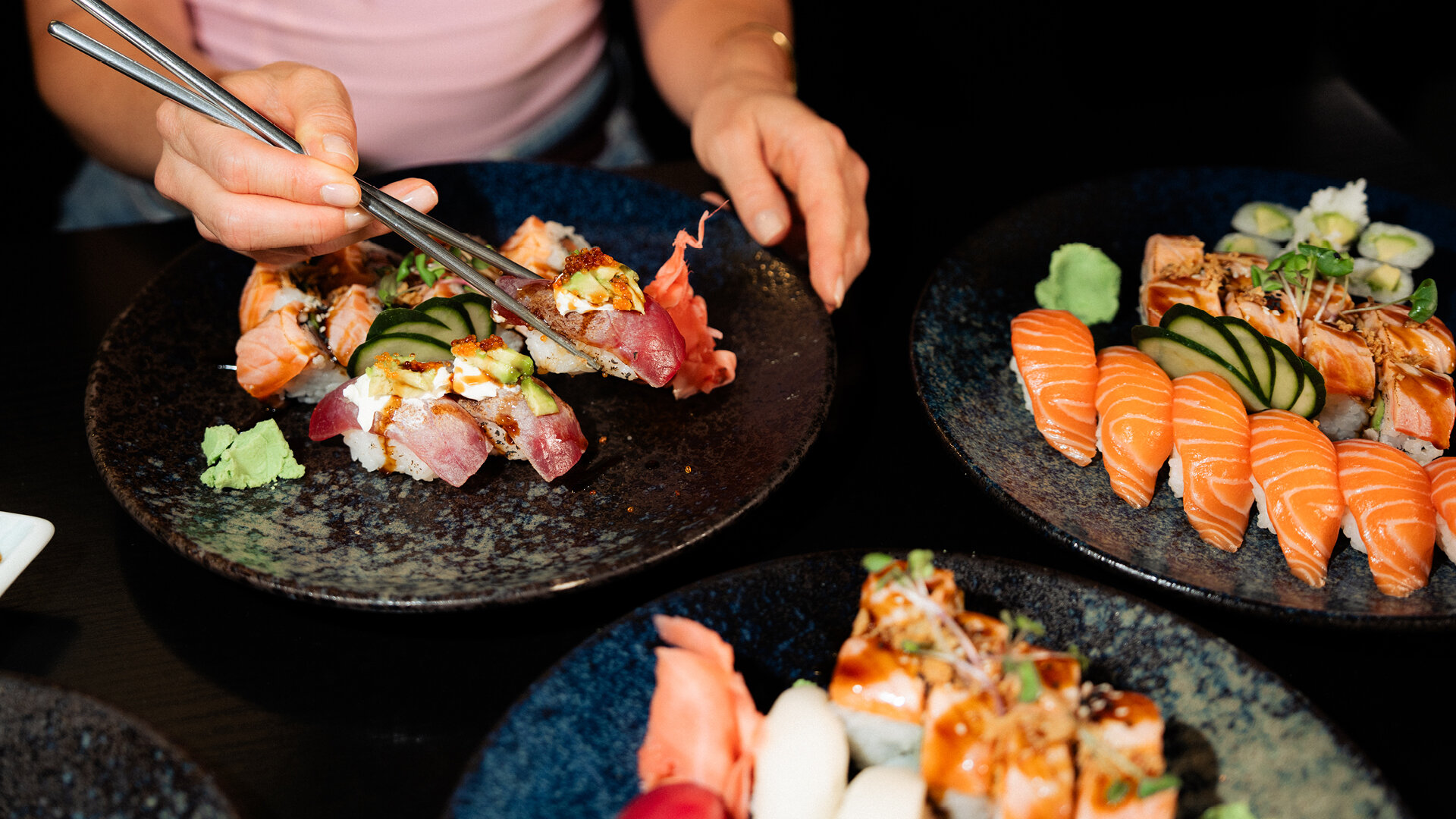Nahaufnahme von Händen die mit silbernen Essstäbchen ein Stück Sushi von dunklen Keramiktellern mit Lachs Nigiri Wasabi und eingelegtem Ingwer aufnehmen.