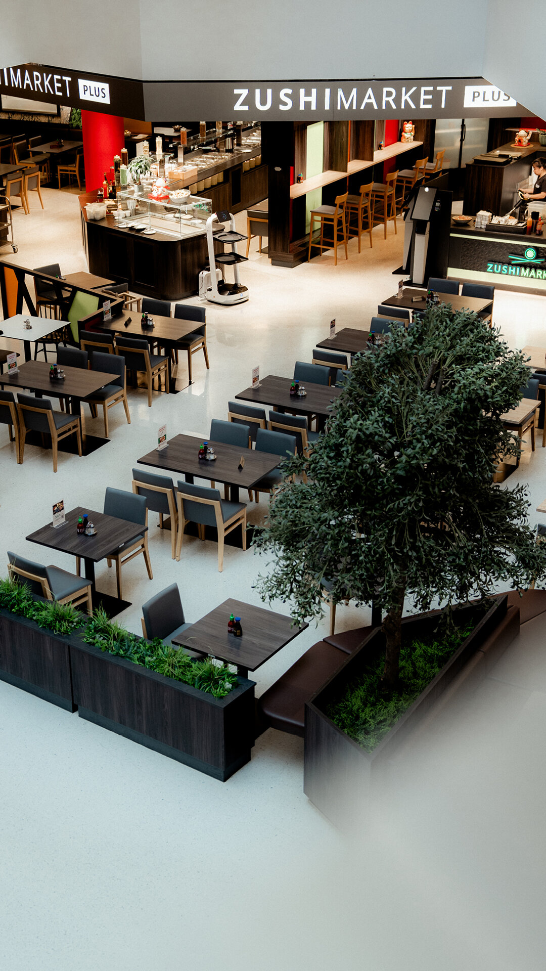 Blick von oben auf den Sitzbereich des Zushimarket Plus Restaurants mit dunkelbraunen Holztischen grauen Stühlen einem großen Baum und Pflanzenkübeln.
