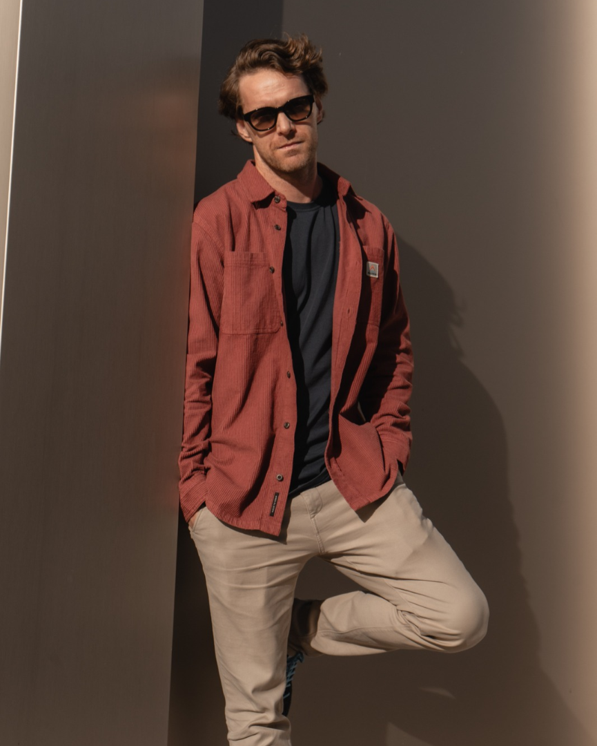 Ein Mann mit Sonnenbrille lehnt in einem rostbraunen Cordhemd, dunklem T-Shirt und beiger Hose an einer modernen Säule im Kaufhaus Tyrol.   