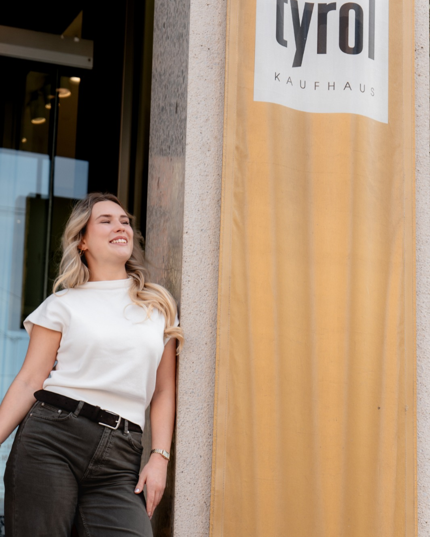Lächelnde junge Frau mit blonden Haaren in weißem T-Shirt und dunkler Jeans lehnt an einer Säule mit Kaufhaus-Tyrol-Banner.   