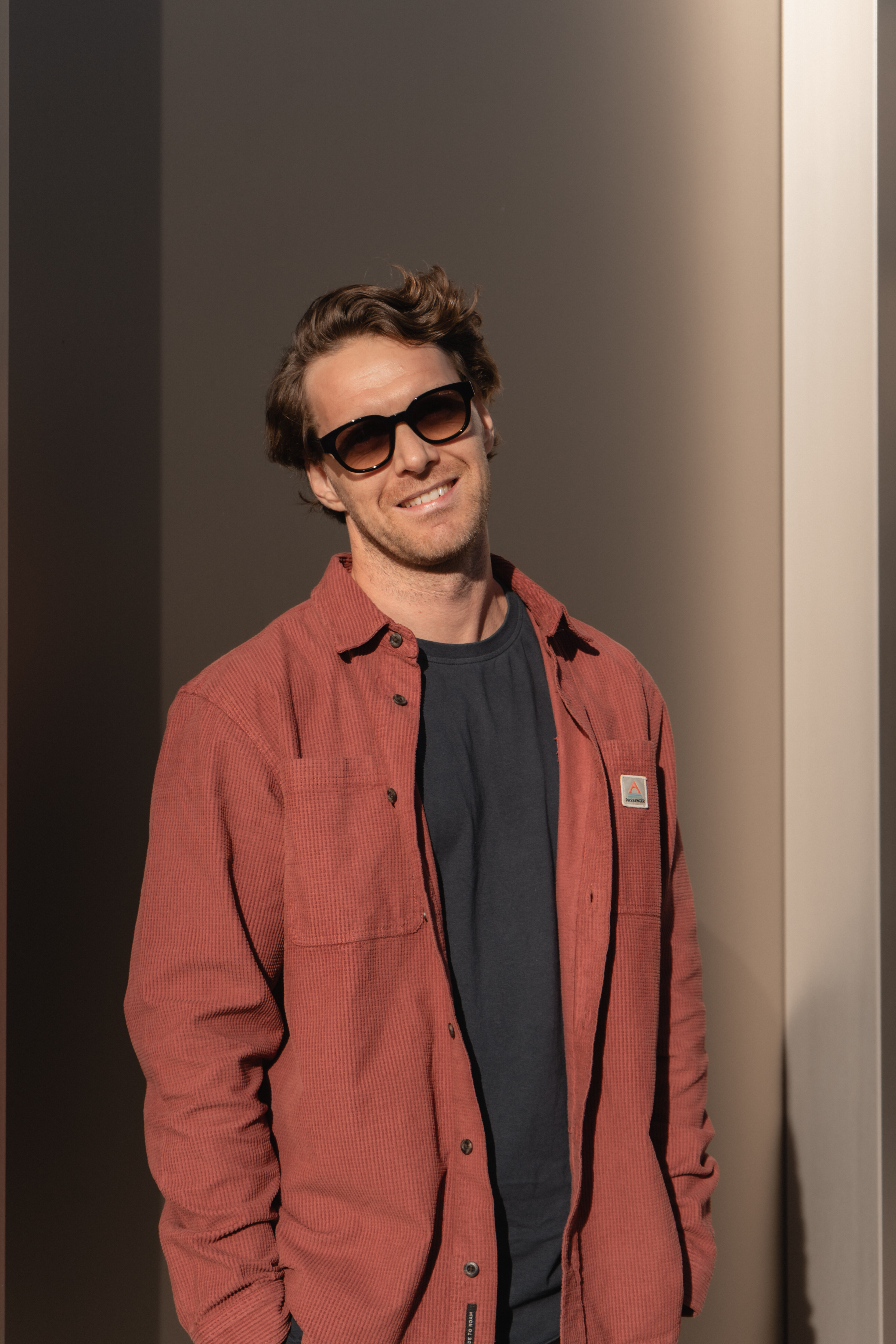 Portrait eines lächelnden Mannes mit braunen Haaren und Sonnenbrille im rostbraunen Cordhemd vor einer modernen Wandfläche.