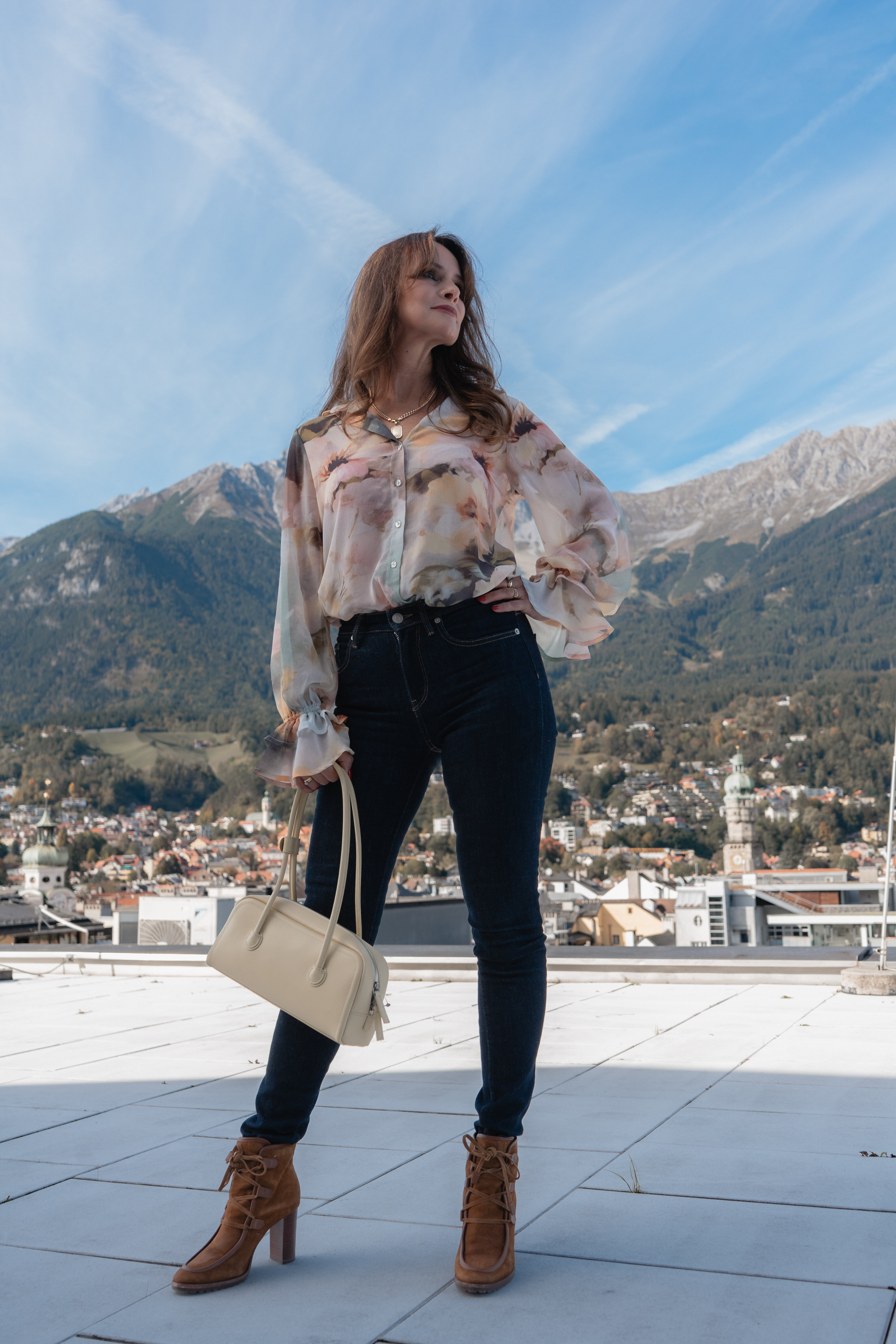 Frau in gemusterter Chiffonbluse, dunkler Skinny Jeans und Stiefeletten steht mit Handtasche auf dem Dach vor dem Innsbrucker Bergpanorama.