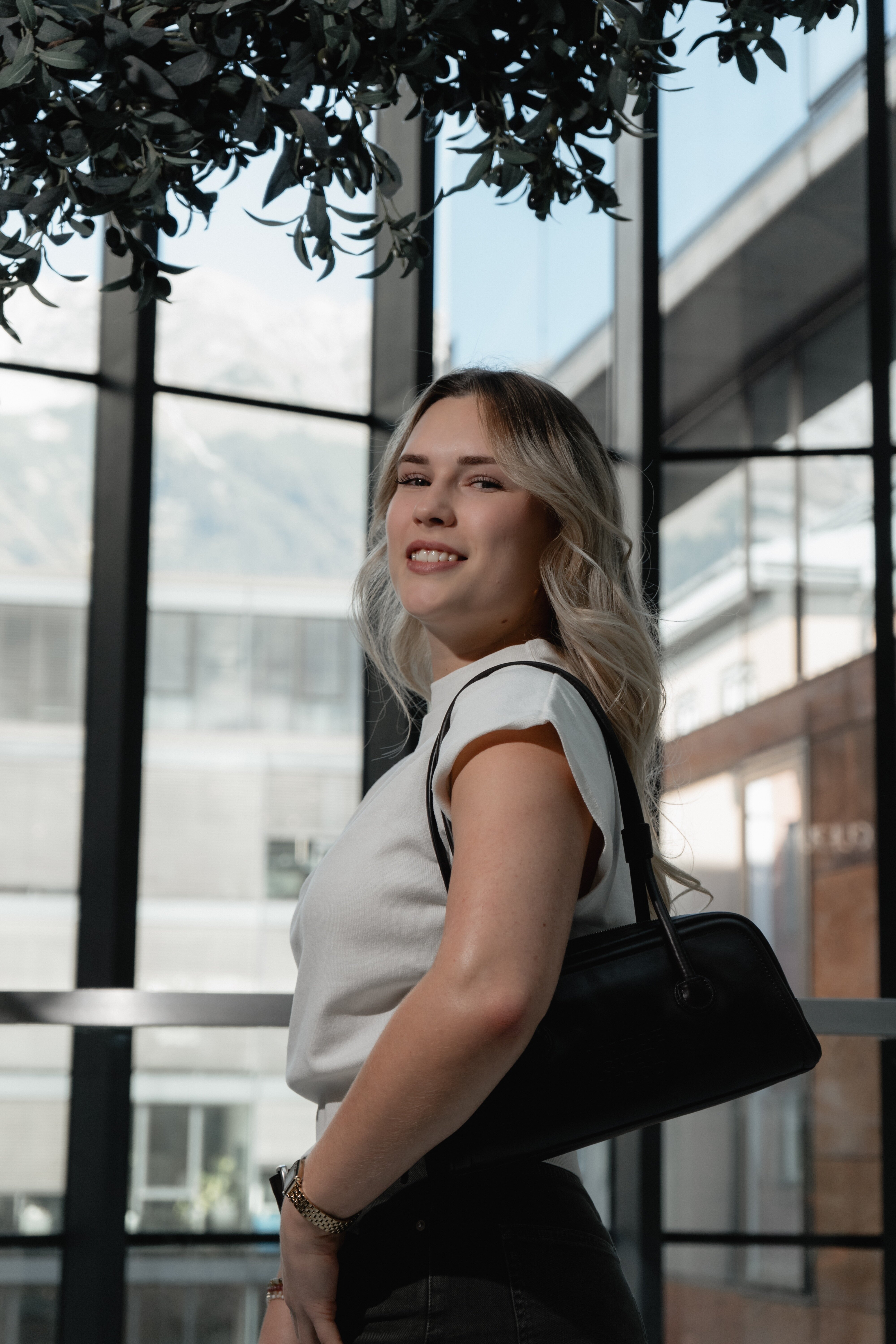 Lächelnde blonde Frau mit Schultertasche und weißem Oberteil blickt unter der modernen Glaskonstruktion des Shoppingcenters in die Kamera.