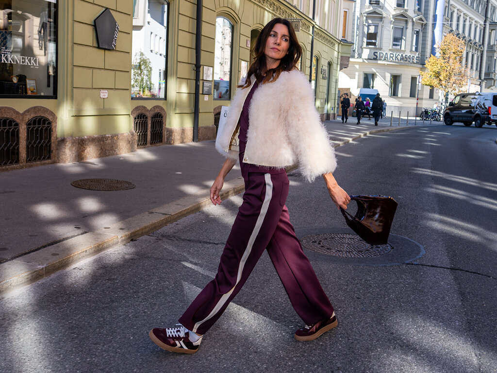 Frau geht in bordeauxfarbener Hose, Sneakern und einer hellen Plüschjacke durch eine Straße in Innsbruck.   