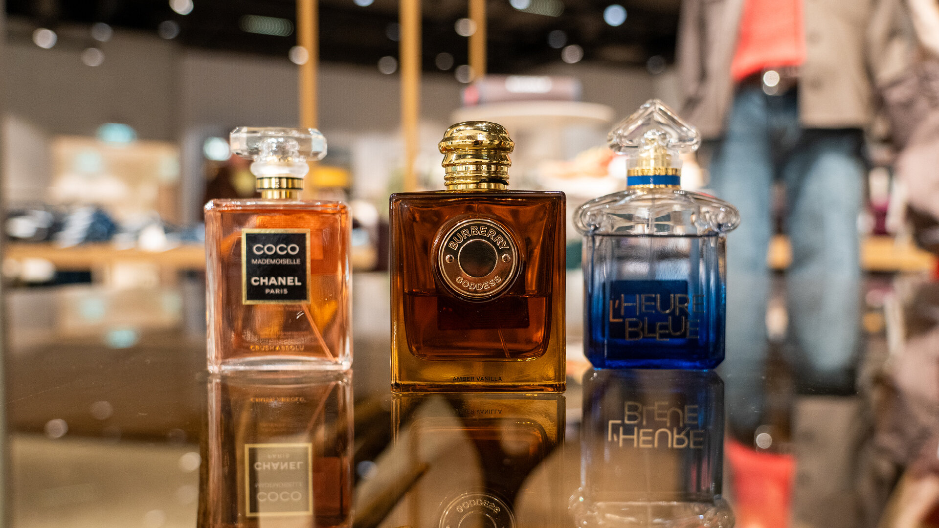 Drei edle Parfüm-Flakons von Chanel, Burberry und Guerlain auf einer spiegelnden Oberfläche nebeneinander aufgestellt.