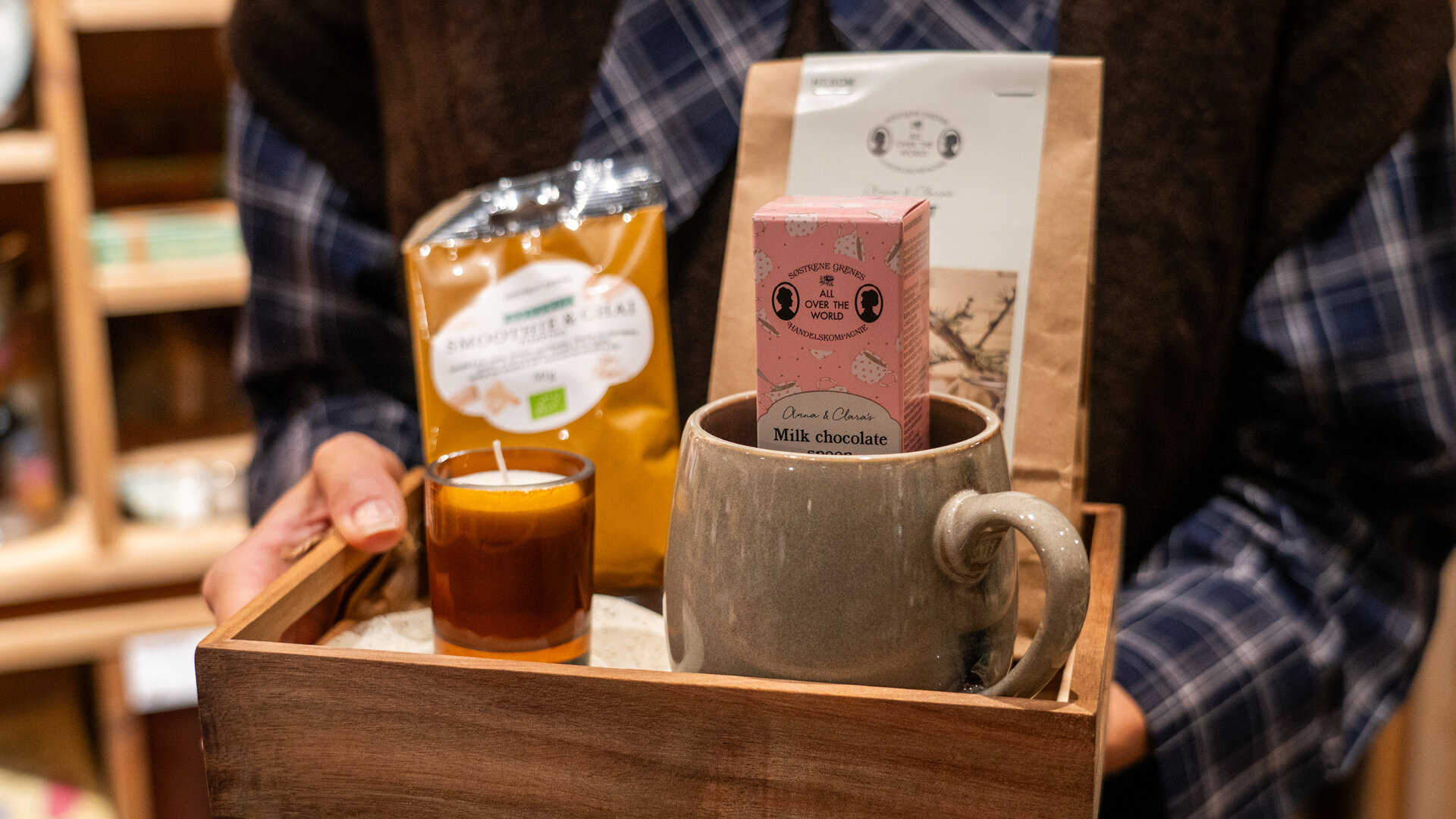 Hände halten ein Holztablett mit einer Steinguttasse, Schokolade, Tee und einer Duftkerze.