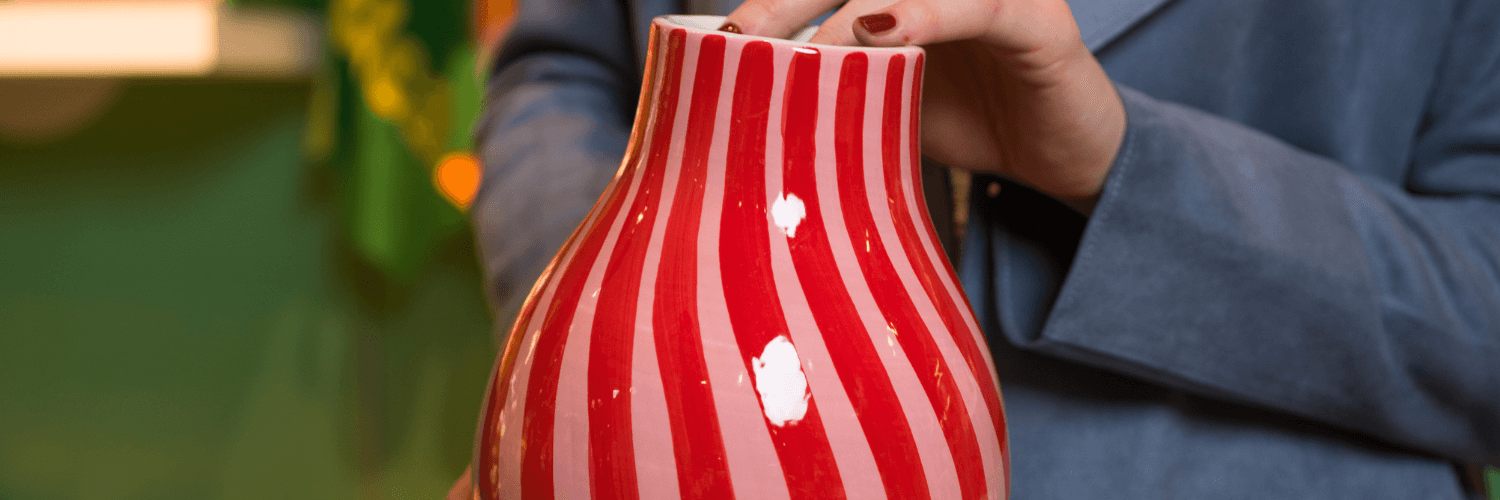 Detailaufnahme einer rot-rosa gestreiften Keramikvase, die von einer Hand sanft berührt wird.