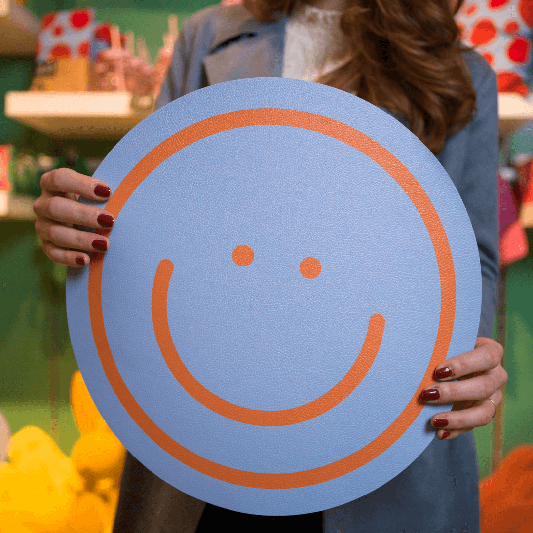 Hände präsentieren ein rundes, hellblaues Tischset mit einem großen, orangefarbenen Smiley-Gesicht.