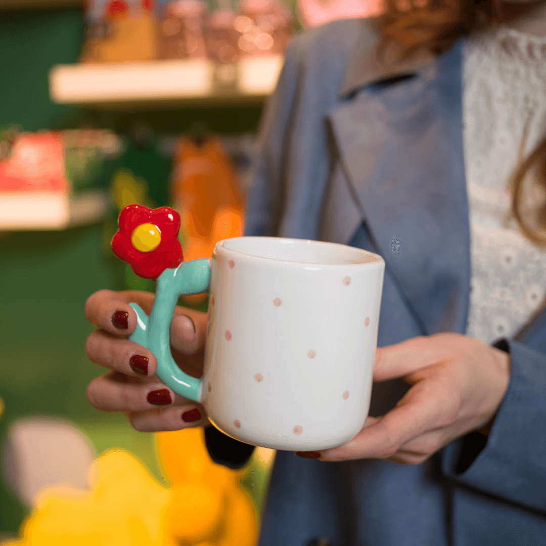 Eine weiße Teetasse mit rosa Punkten und einem Henkel, der wie ein Blumenstiel mit roter Blüte geformt ist.