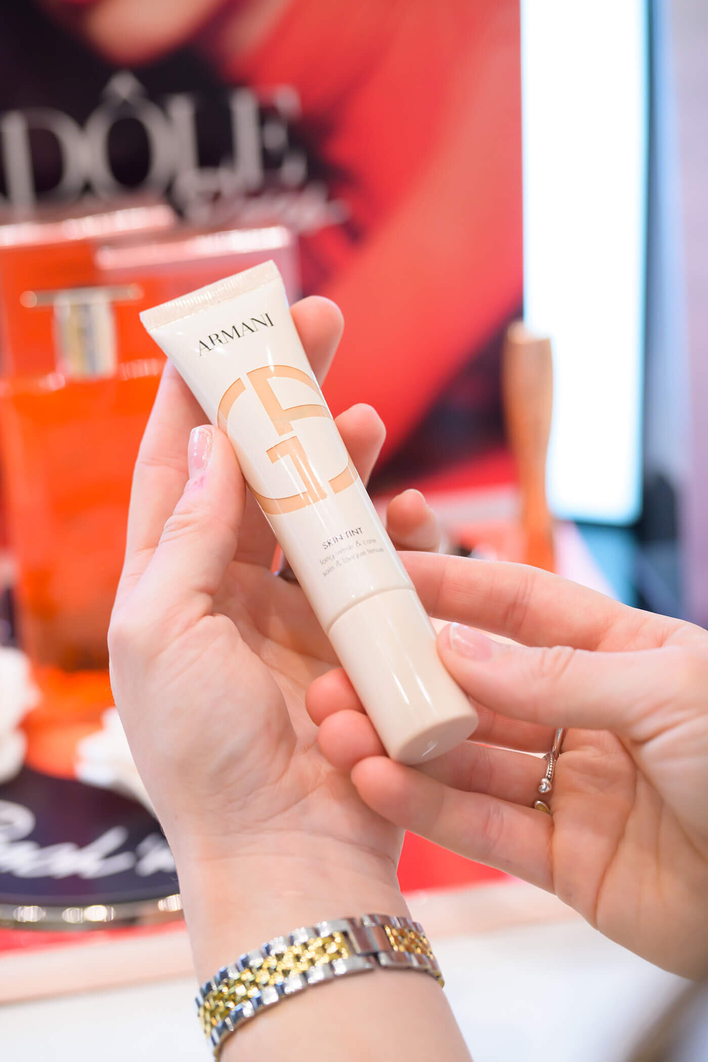 Zwei Hände halten eine hellbeige Tube des „Giorgio Armani Skin Tint“ Make-ups vor einem verschwommenen, roten Hintergrund.