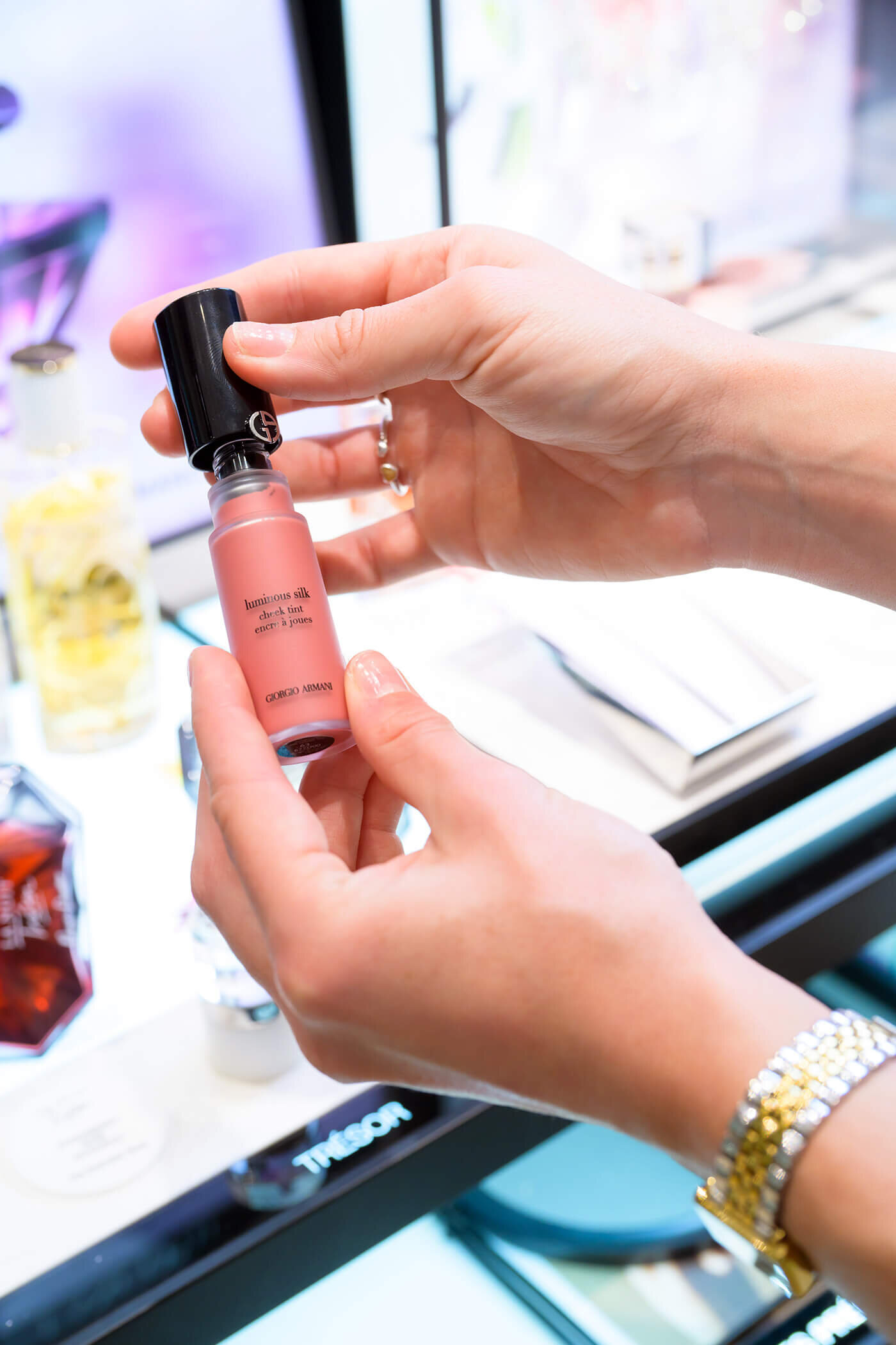 Ein Fläschchen des flüssigen „Giorgio Armani Luminous Silk Cheek Tint“ Rouge in einem zarten Rosatun wird von zwei Händen präsentiert.