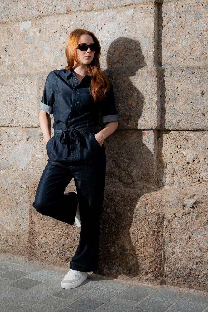 Eine Frau mit Sonnenbrille und dunkelblauem Denim-Jumpsuit lehnt an einer hellen Natursteinwand.