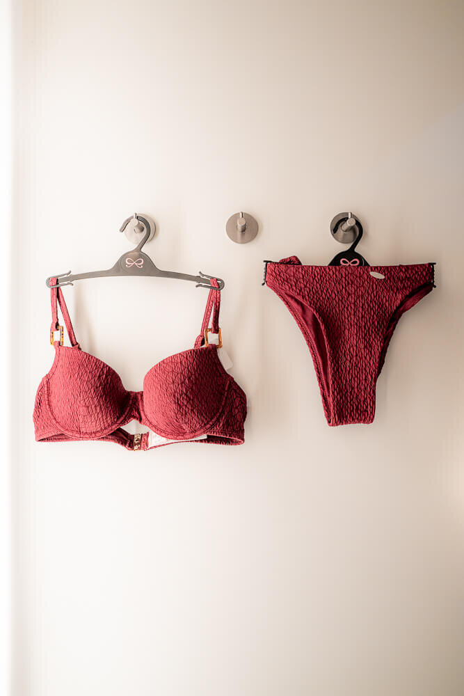 Ein dunkelroter Bikini mit strukturierter Oberfläche hängt auf zwei Kleiderbügeln an Wandhaken in einer Umkleidekabine.
