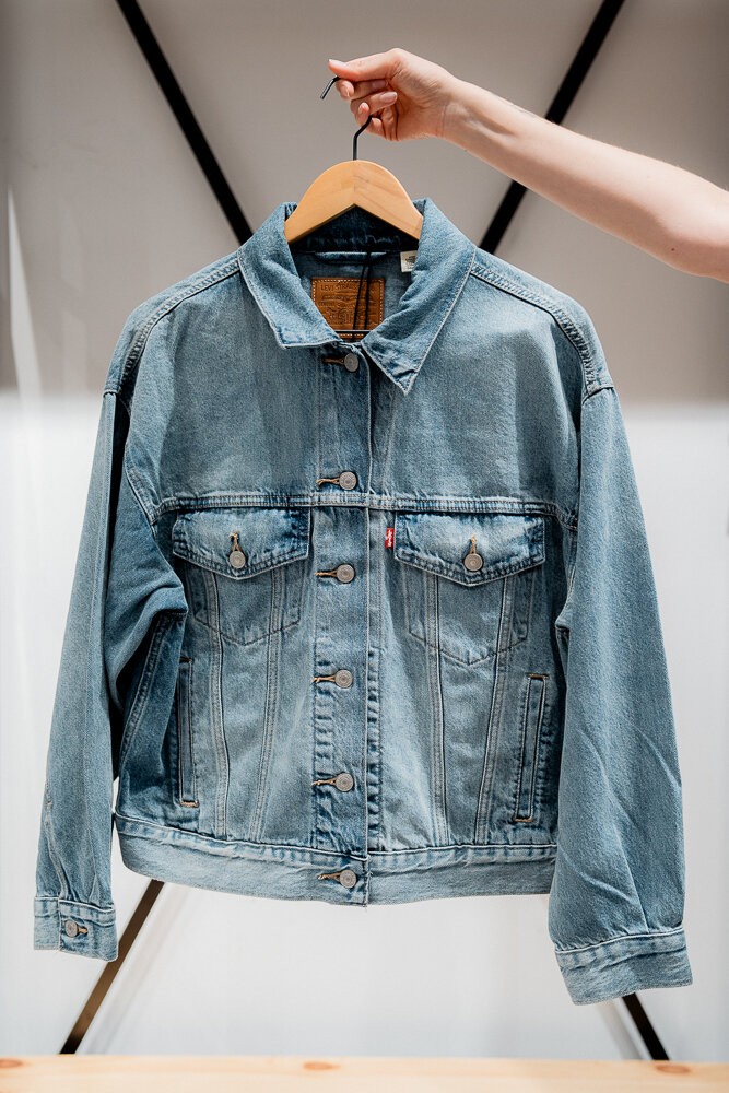 Eine Hand hält eine klassische blaue Denim-Jeansjacke der Marke Levi's auf einem Holzkleiderbügel nach oben.