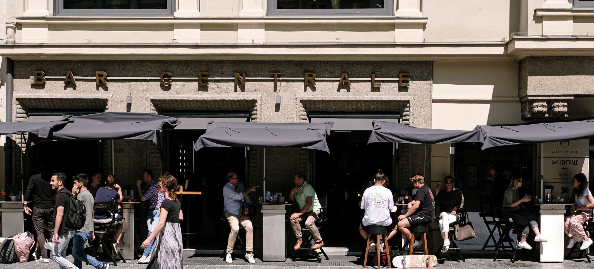 Zahlreiche Gäste sitzen im sonnigen Außenbereich unter schwarzen Sonnenschirmen vor der Fassade der Bar Centrale.