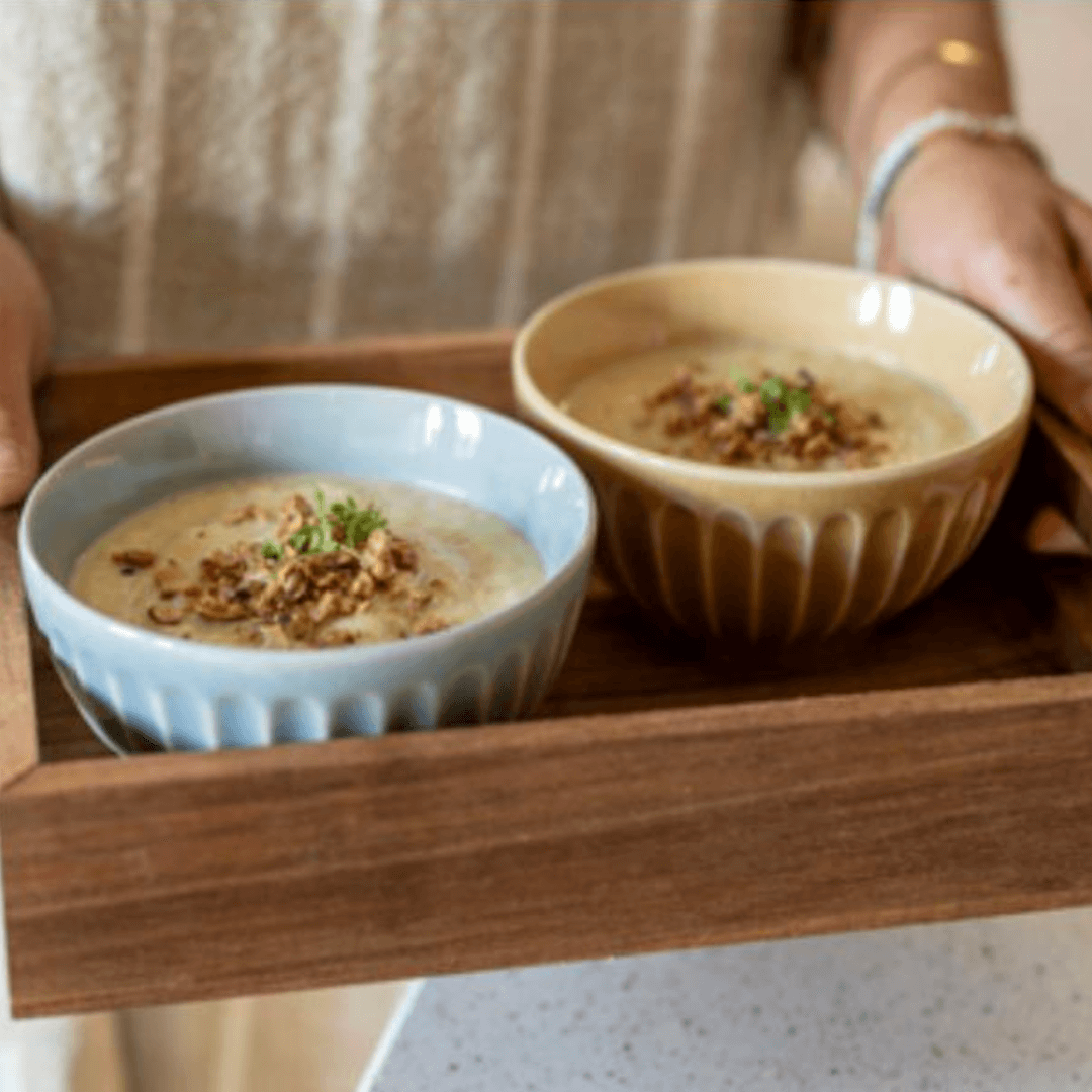Hände halten ein Holztablett mit zwei Schüsseln cremiger Birnen-Sellerie-Suppe, getoppt mit Knusper-Topping und frischen Kräutern.