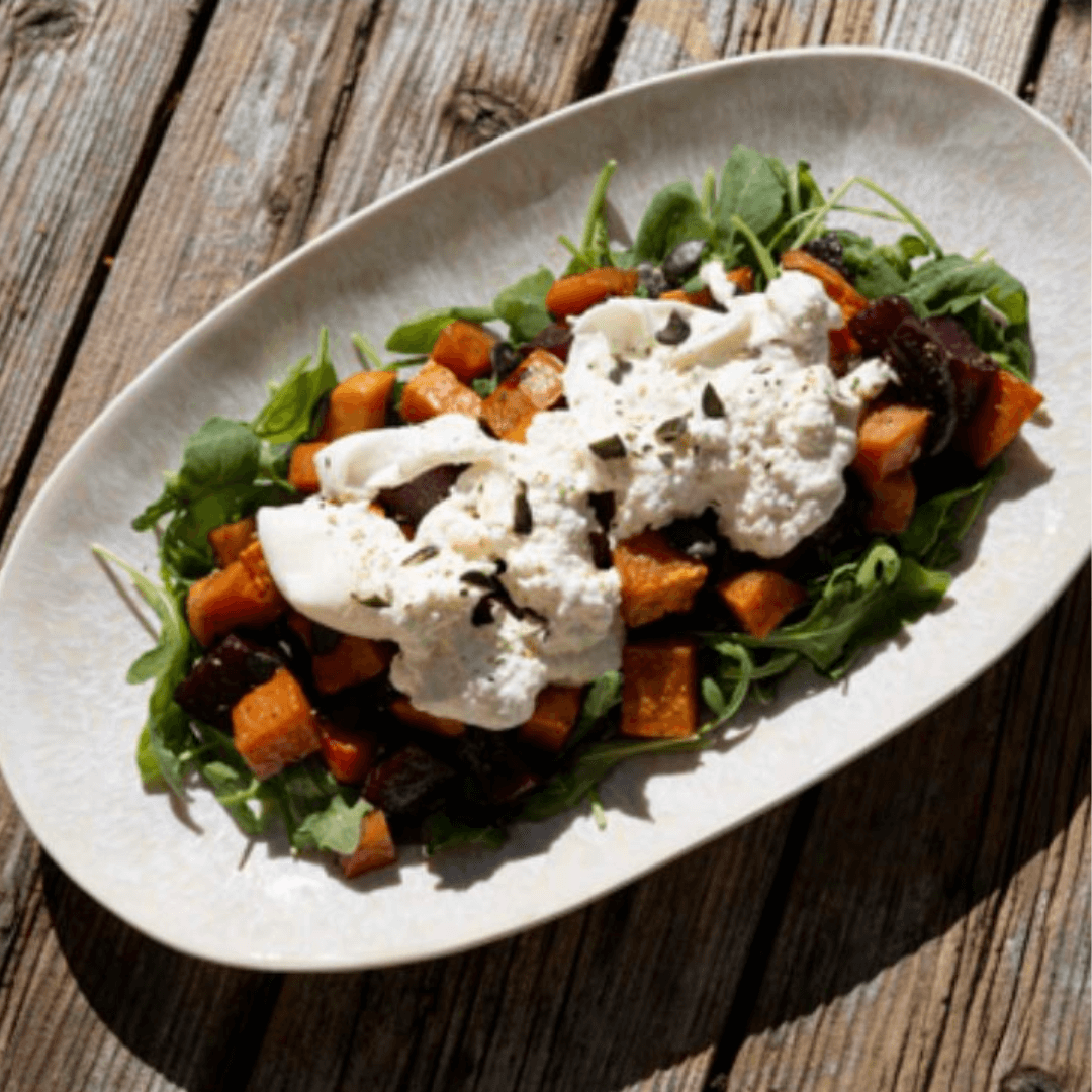 Ein ovaler Teller mit Röstgemüse-Salat, bestehend aus Rucola, geröstetem Kürbis, Roter Bete und einer großen Kugel cremiger Burrata, bestreut mit Kernen.