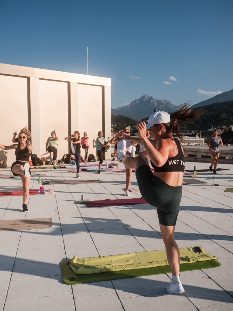 Eine Fitness-Gruppe führt dynamische Kick-Übungen während eines Zirkeltrainings auf der sonnigen Dachterrasse in Innsbruck aus.