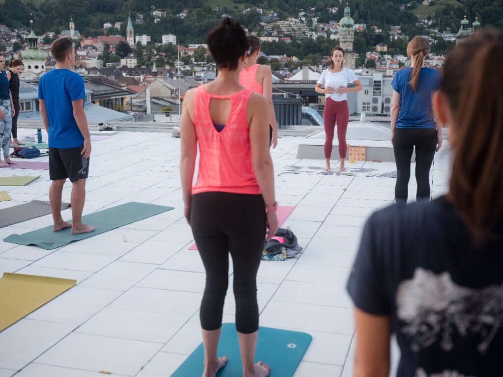 Eine Pilates-Gruppe steht auf Gymnastikmatten auf einer Dachterrasse, während die Trainerin im Hintergrund Atemübungen vor der Innsbrucker Kulisse anleitet.