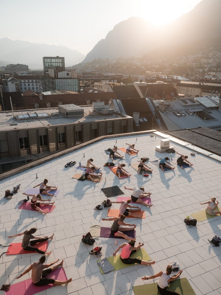 Vogelperspektive einer Yoga-Gruppe auf bunten Matten bei Dehnübungen auf der Dachterrasse des Kaufhaus Tyrol im Abendlicht.