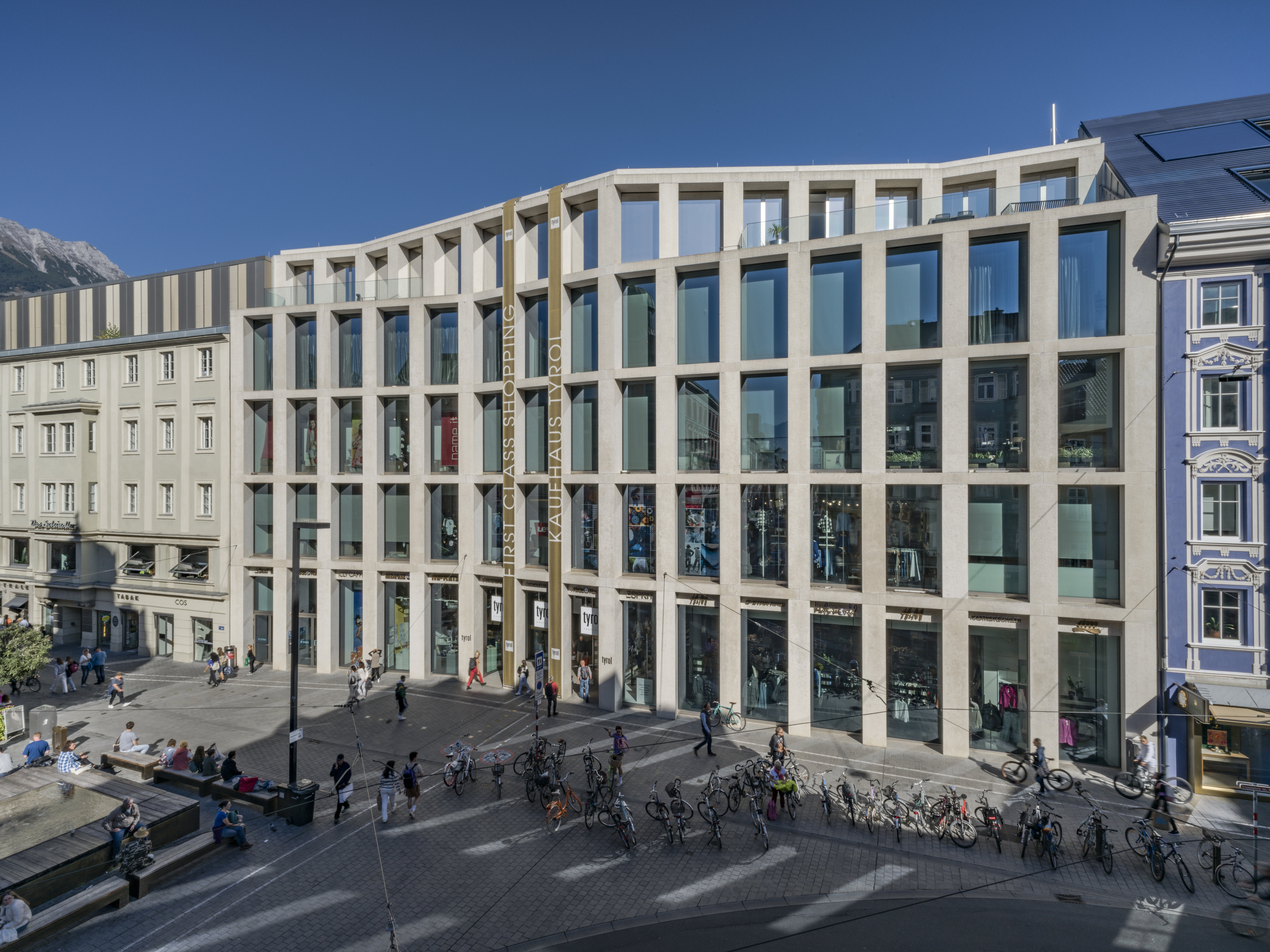 Die helle, moderne Beton- und Glasfassade des Kaufhaus Tyrol an der Fußgängerzone in der Maria-Theresien-Straße mit Passanten und Fahrrädern davor.