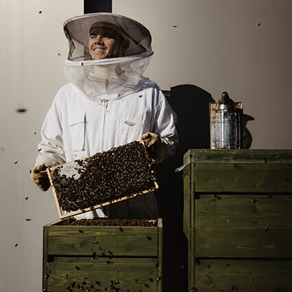Ein lächelnder Imker in weißer Schutzkleidung hält eine Bienenwabe neben seinen Bienenstöcken.