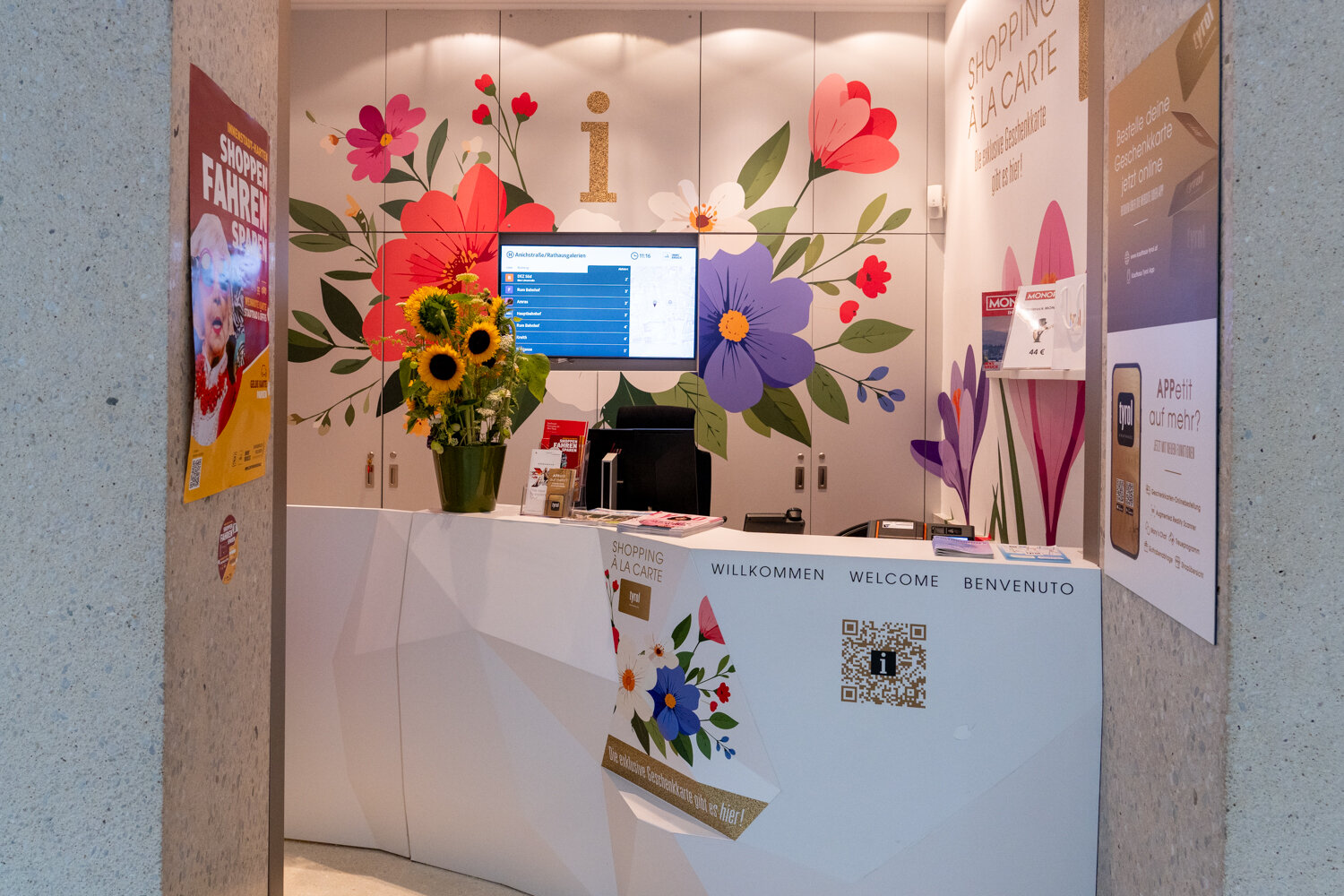 Moderner, weißer Informationstresen im Kaufhaus Tyrol mit bunten Blumenillustrationen an der Rückwand, einem Infoscreen und einem Strauß Sonnenblumen.