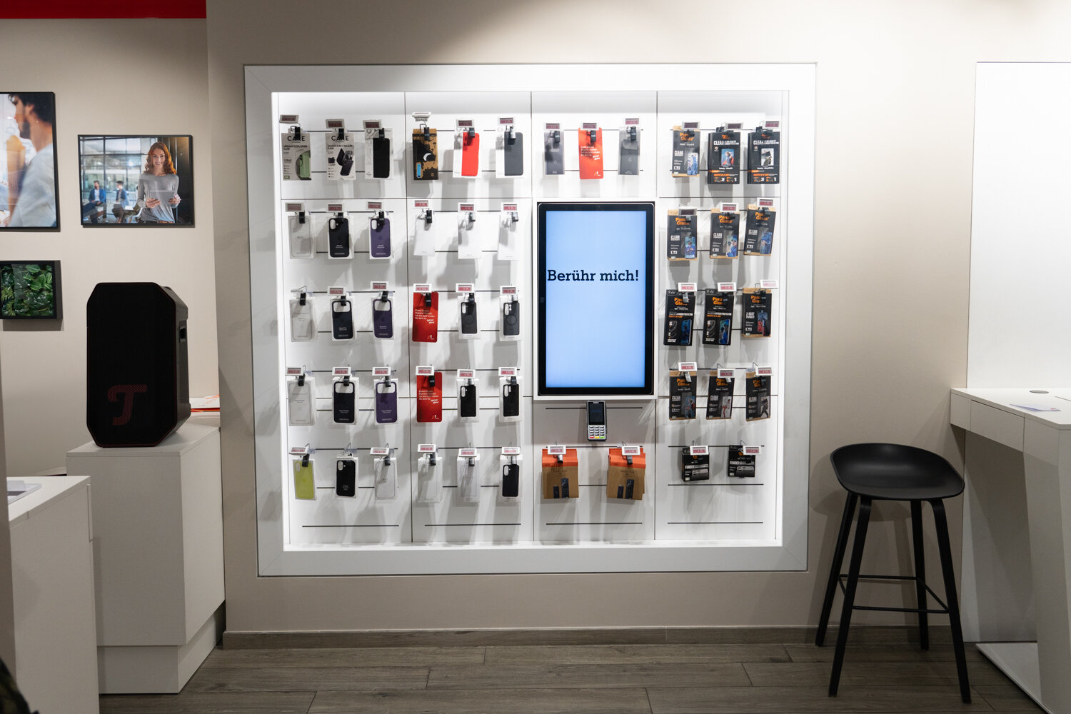 Beleuchtetes Wandregal im A1 Shop mit Smartphone-Hüllen, Zubehör und einem interaktiven Display mit der Aufschrift „Berühr mich!“.