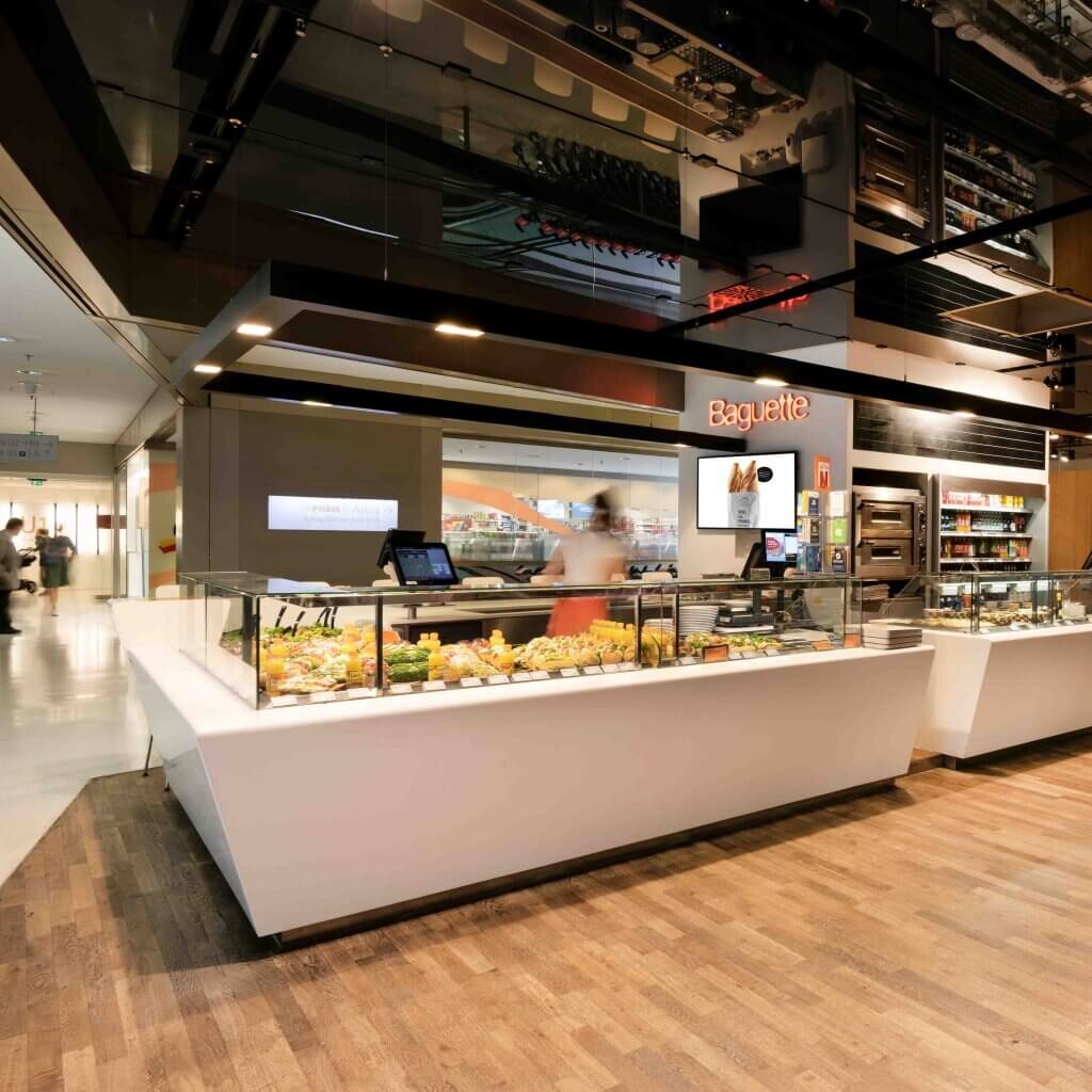 Ein moderner, hell erleuchteter Verkaufstresen einer Baguette-Bäckerei mit einer großen Auswahl an frisch belegten Snacks, Sandwiches und Salaten in der Glasvitrine. Im Hintergrund ist das beleuchtete orangefarbene Baguette-Logo zu sehen. Der Laden zeichnet sich durch ein zeitgemäßes Design mit Holzboden und minimalistischen weißen Thekenelementen aus.