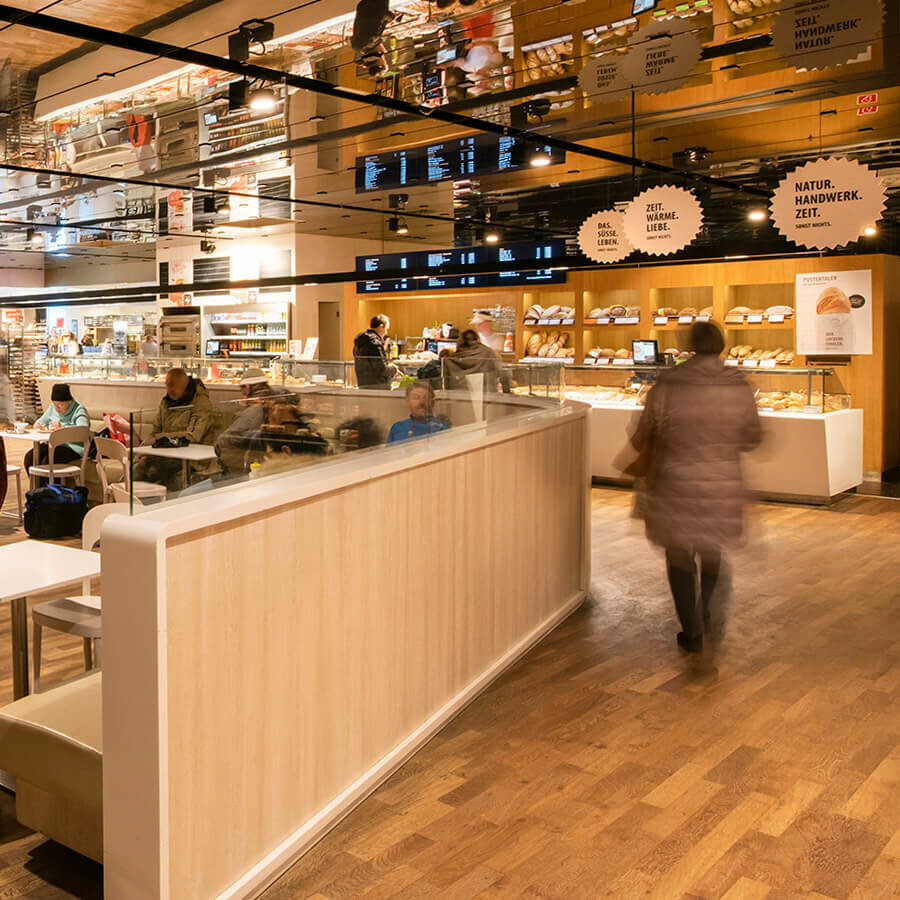Innenansicht einer Baguette-Filiale mit Blick auf den Café-Sitzbereich auf der linken Seite und die Brottheke im Hintergrund. Kunden bewegen sich durch den großzügigen Verkaufsraum mit stilvollem Holzboden. An der Decke befinden sich dekorative, runde Schilder mit Aufschriften wie „Natur. Handwerk. Zeit.“ und die Deckenoberfläche spiegelt den hellen Raum wider.