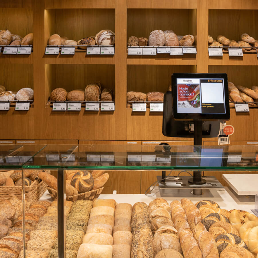 Detailaufnahme der Brottheke und der Holzregale in einer Baguette-Bäckerei. Die Regale sind prall gefüllt mit verschiedenen frisch gebackenen Brotsorten, Handsemmeln und Körnerbrötchen. Im Vordergrund steht ein digitaler Kassenbildschirm zur Produkterfassung direkt an der gläsernen Verkaufsvitrine.