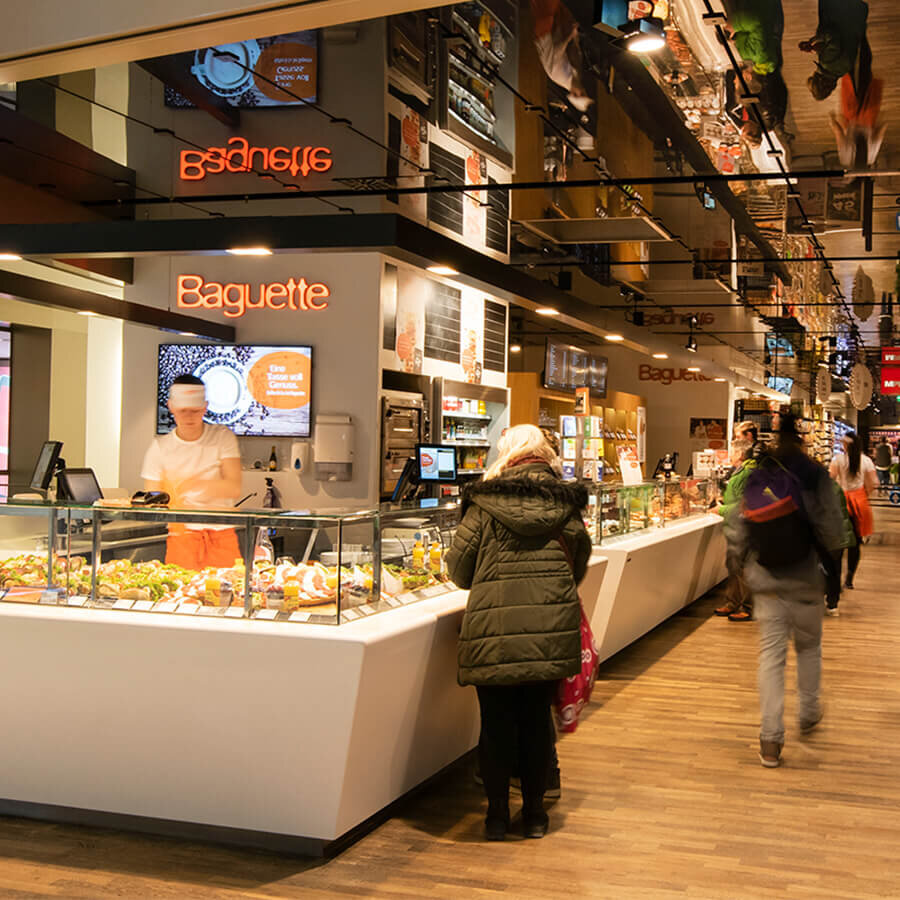 Eine Kundin steht am modernen, weißen Verkaufstresen einer Baguette-Filiale, während ein Mitarbeiter hinter der Vitrine frische Snacks zubereitet. Über der Theke leuchtet das orangefarbene Markenlogo neben einem Bildschirm, der Kaffeespezialitäten bewirbt. Der Verkaufsraum besticht durch warmen Holzboden und eine markante, spiegelnde Deckenoptik.