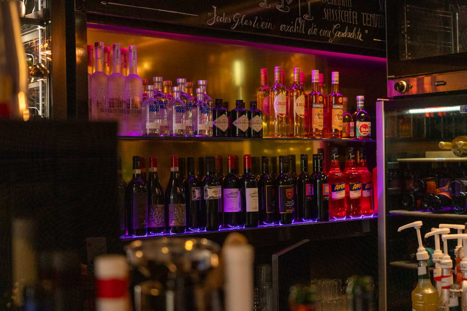 Bunt beleuchtete Regale mit Wodka-, Gin-, Aperol- und Weinflaschen an einer Bar.