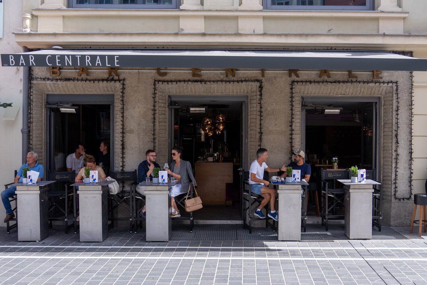 Gäste sitzen an kleinen Stehtischen vor der steinernen Fassade der Bar Centrale unter einer dunklen Markise.