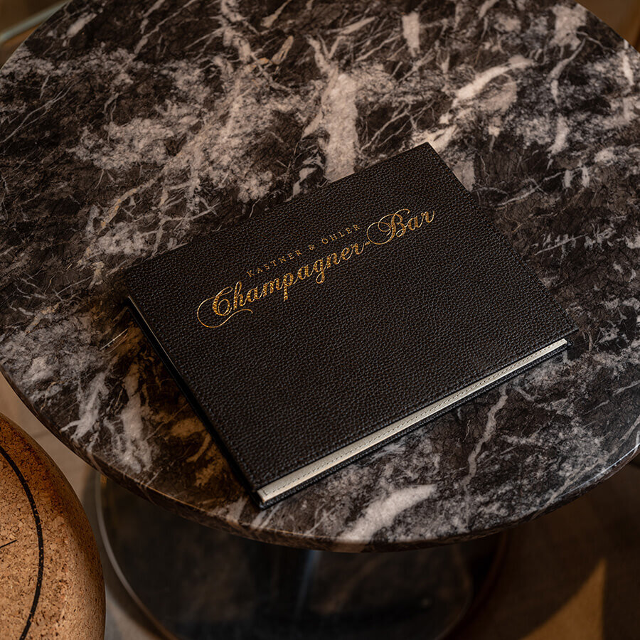 Schwarze Lederkarte mit goldener Aufschrift Kastner & Öhler Champagner-Bar auf einem rundem Marmortisch.