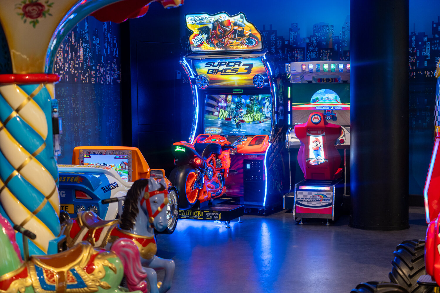 Verschiedene Spiel- und Fahr-Automaten wie Mario Kart Arcade DX und Super Bikes unter blauem Licht.