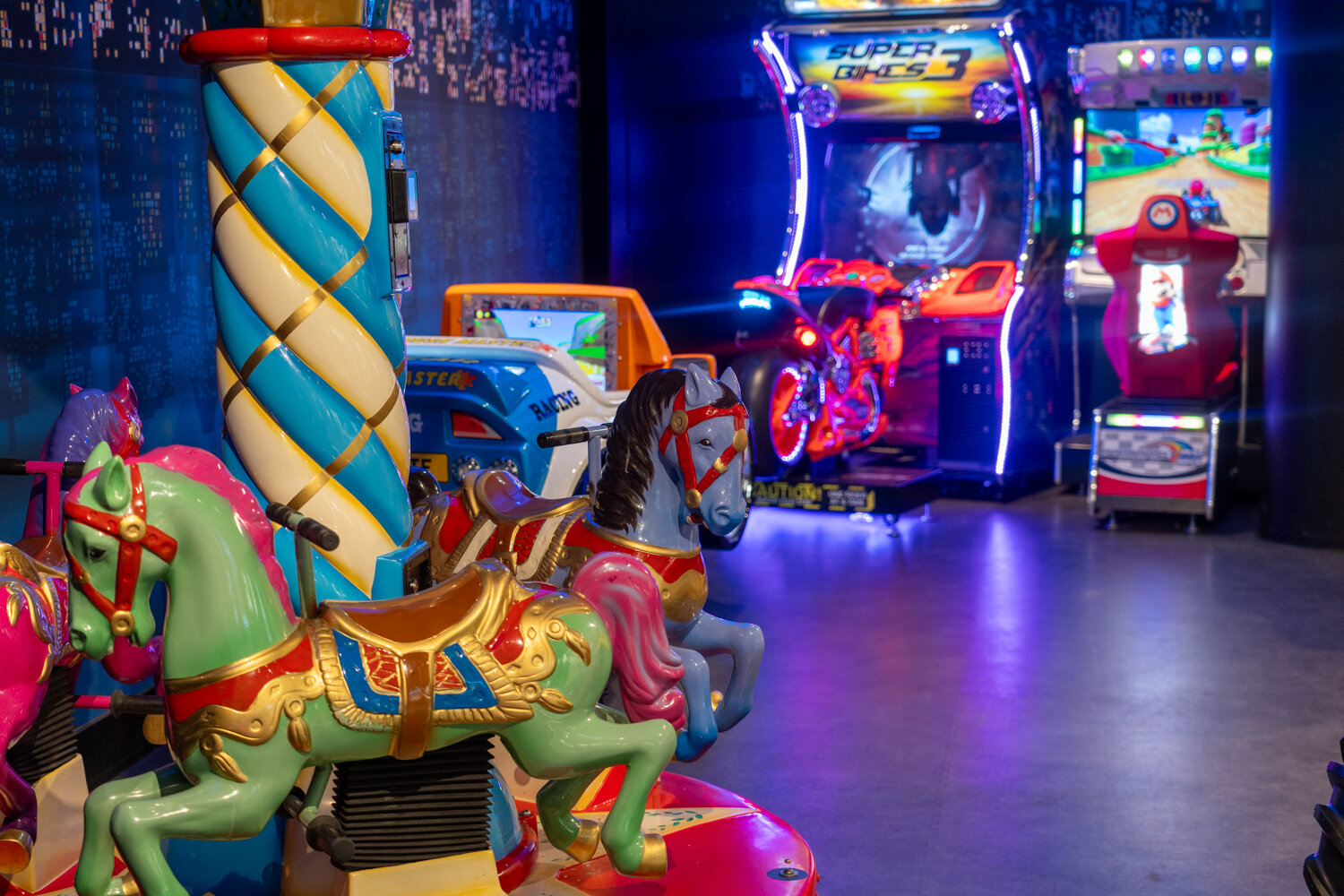 Bunte Karussell-Pferde im Vordergrund mit Fahr-Simulatoren und Arcade-Spielen im Hintergrund.