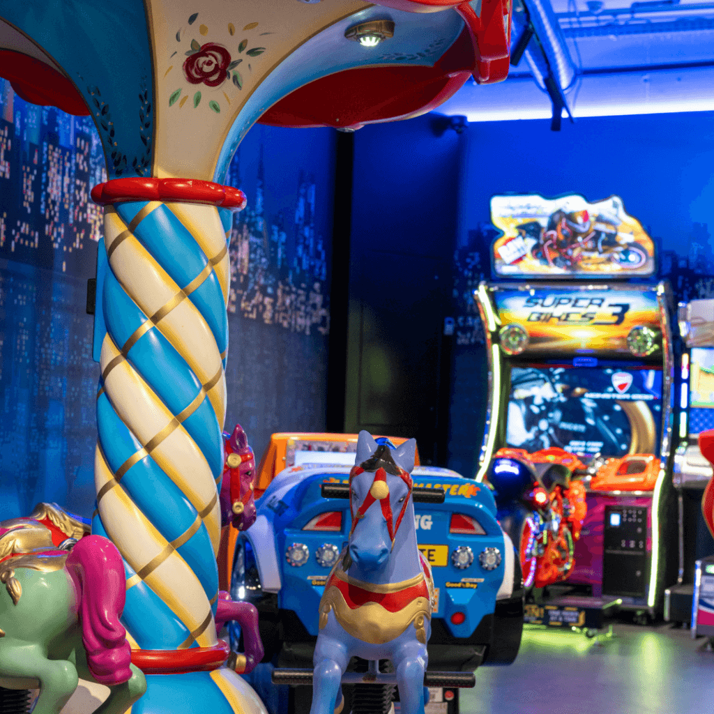 Ein buntes Kinderkarussell mit Pferden im Vordergrund und leuchtende Arcade-Rennspielautomaten im Hintergrund.