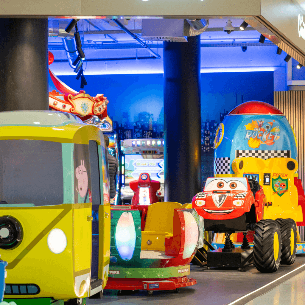 Kinderbereich mit verschiedenen bunten Fahrgeschäften wie einem Peppa-Wutz-Bus, einem roten Rennauto und einer Spielzeug-Rakete.